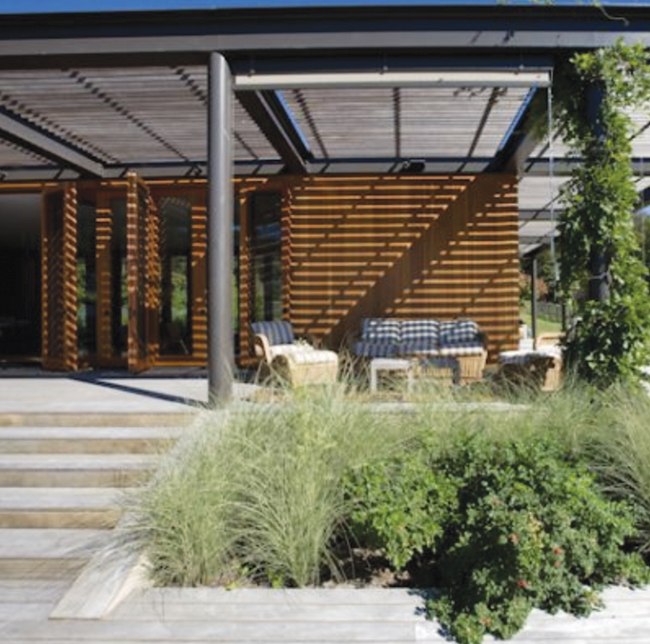 A modern outdoor living space with a pergola-covered patio, wooden slat accents, and lush greenery.