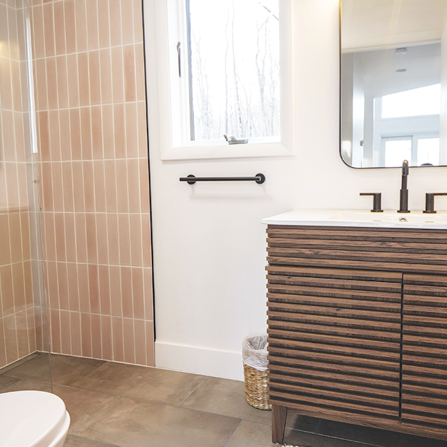 A modern bathroom with a wood-slatted vanity, black fixtures, soft pink vertical tiles, and a sleek mirror.