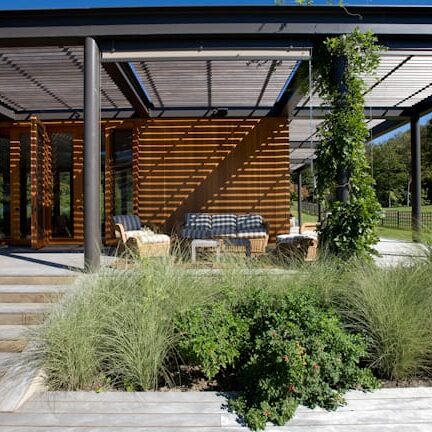 New Construction A modern outdoor living space with a pergola-covered patio, wooden slat accents, and lush greenery.