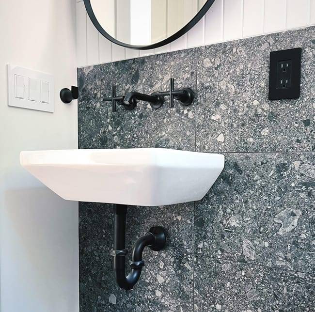 Wall-mounted white sink with black hardware against a dark gray stone tile backdrop